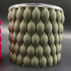 Puede incluir: Un recipiente cilíndrico con un exterior texturizado verde oliva y una parte superior transparente y redondeada. El exterior presenta un patrón repetitivo de formas redondeadas en forma de escamas. El recipiente es probablemente de plástico o un material similar.