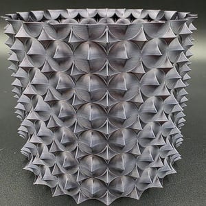 May include: A gray, 3D-printed planter with a textured surface. The planter has a geometric design with repeating circular and triangular shapes, creating a spiky appearance. The planter is approximately 6 inches tall and 6 inches wide.