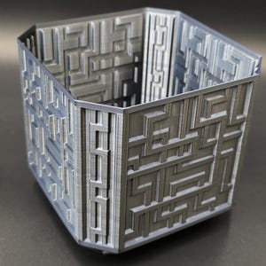 Puede incluir: Un recipiente hexagonal gris con un patrón de laberinto en su exterior. Las paredes están intrincadamente diseñadas con una serie de líneas interconectadas, creando una apariencia de laberinto complejo. El recipiente está hecho de un material sólido y tiene un acabado liso.