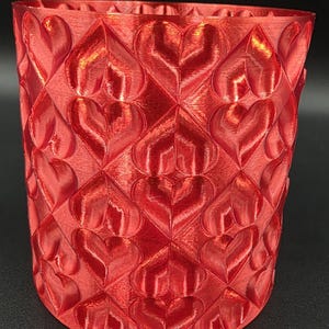 May include: A red, heart-patterned vase. The vase has a repeating heart design across its surface. The hearts are raised, creating a textured appearance. The vase is cylindrical with a slightly flared top, and is made of a shiny, reflective material.