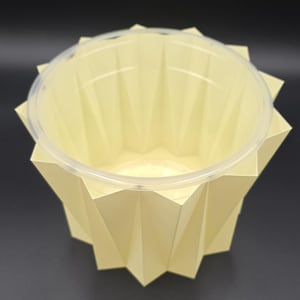 Puede incluir: Un recipiente de estilo origami amarillo claro con una tapa de plástico transparente. El recipiente tiene un diseño geométrico en forma de estrella. El recipiente está vacío y listo para ser llenado.