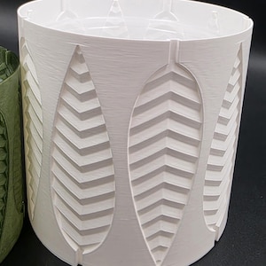May include: A white cylindrical planter with a textured surface and leaf-shaped cutouts. The cutouts have a chevron pattern. The planter is made of a hard material and is designed for indoor or outdoor use.