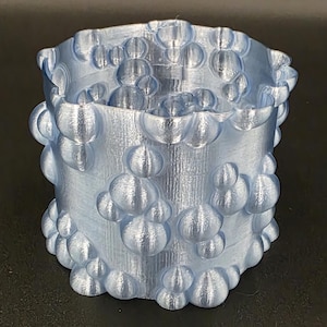 Puede incluir: Un recipiente cilíndrico azul claro con un patrón texturizado en forma de burbujas. El recipiente tiene una parte superior ligeramente ensanchada y está hecho de un material brillante de aspecto metálico. Las burbujas varían en tamaño.