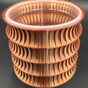Puede incluir: Un recipiente decorativo de color cobre, impreso en 3D, con un borde de plástico transparente. El recipiente tiene un diseño acanalado y en capas, creando una apariencia texturizada. La forma general es cilíndrica, adecuado para guardar objetos pequeños o como pieza decorativa.