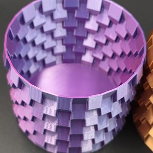 Puede incluir: Un recipiente cilíndrico impreso en 3D con un patrón tejido en tonos de morado y lavanda. El interior es de color lavanda sólido. El recipiente está diseñado con un exterior geométrico texturizado.