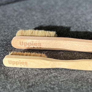 以下が含まれることがあります： 天然毛のブラシ2本。ブラシの柄には「Uppies & co.」の文字がプリントされています。1本は明るい色の木材、もう1本は竹でできています。