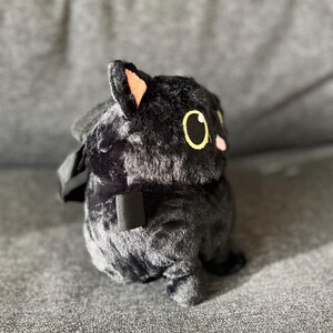 May include: A plush black cat-shaped backpack with yellow eyes and pink cheeks. The cat has a small, black strap and a black handle. The plush toy is sitting on a gray surface.