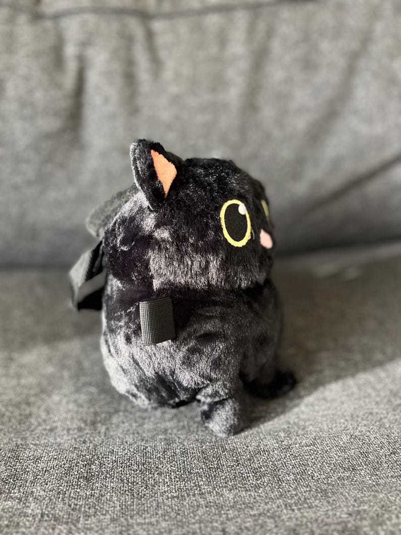 May include: A plush black cat-shaped backpack with yellow eyes and pink cheeks. The cat has a small, black strap and a black handle. The plush toy is sitting on a gray surface.