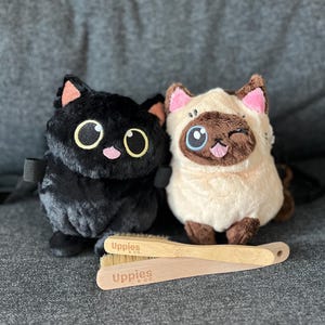 May include: Two plush cat toys, one black and one cream-colored, sit side-by-side. The black cat has large yellow eyes and pink accents. The cream cat winks and has brown and pink details. Two wooden brushes with the text "Uppies & co." are in front.