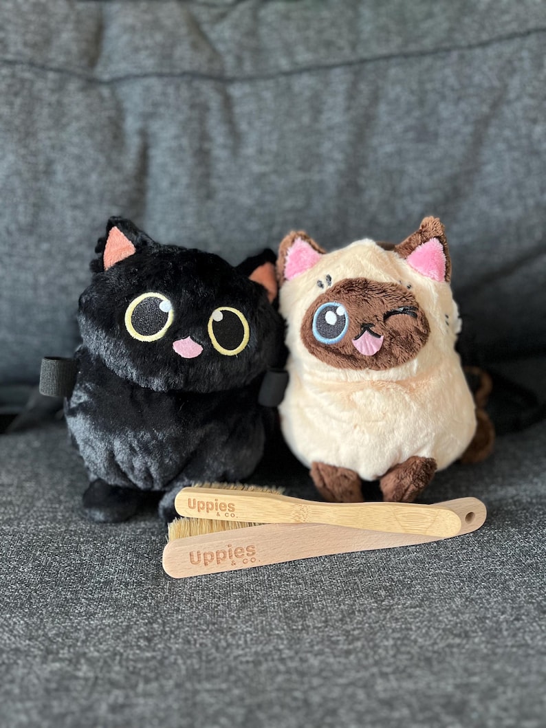 May include: Two plush cat toys, one black and one cream-colored, sit side-by-side. The black cat has large yellow eyes and pink accents. The cream cat winks and has brown and pink details. Two wooden brushes with the text "Uppies & co." are in front.