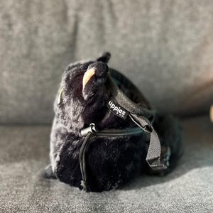 May include: A black, plush bag with a drawstring closure and a small zipper pocket. The bag has a soft, furry texture and a black strap. The brand name "uppies & co." is visible on a small tag.