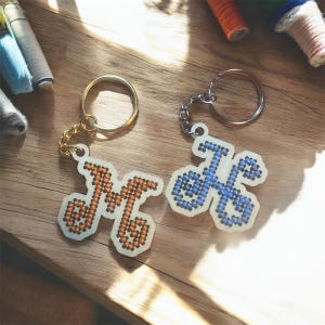 May include: Two letter-shaped keychains featuring a white base with embroidered details. One keychain displays the letter M stitched in orange thread, while the other features the letter H stitched in light blue thread, both with gold-tone rings.