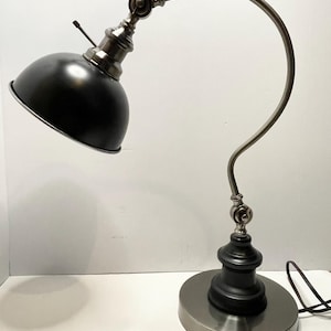 May include: A black and silver desk lamp with an adjustable curved arm and a round base. The lamp has a black lampshade and a silver-toned metal finish, with a black power cord.