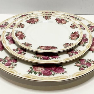 May include: A set of three white ceramic plates with a floral rose design. The plates feature a gold trim and a repeating pattern of pink and red roses with green leaves. The plates are stacked on top of each other.