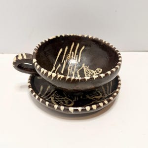 May include: A dark brown ceramic teacup and saucer set. The cup has a handle and is decorated with white painted designs. The rim of the cup and saucer are trimmed with white. The set is likely handmade and suitable for serving tea or coffee.