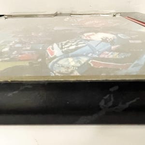 May include: A black framed box with a picture of a race car driver. The image is slightly blurred, but the driver is wearing a blue and red racing suit. The box has a silver hinge and a red accent on the side.