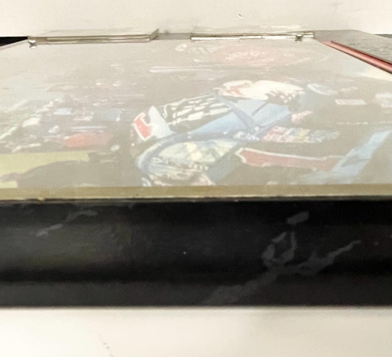 May include: A black framed box with a picture of a race car driver. The image is slightly blurred, but the driver is wearing a blue and red racing suit. The box has a silver hinge and a red accent on the side.