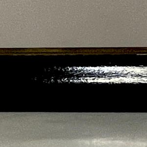May include: A long, black, rectangular object with a shiny surface. A thin, brown, metal piece is attached to the top, with a small, dark knob at one end. The object is set against a light gray background.