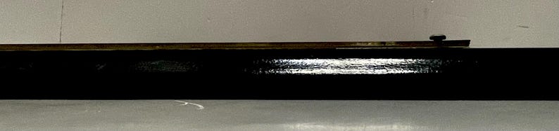 May include: A long, black, rectangular object with a shiny surface. A thin, brown, metal piece is attached to the top, with a small, dark knob at one end. The object is set against a light gray background.