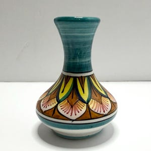 May include: A ceramic vase with a narrow neck and a wide, rounded base. The neck is painted in horizontal teal bands. The body features a repeating petal pattern in yellow, brown, and pink, outlined in black, with teal and white bands at the base.
