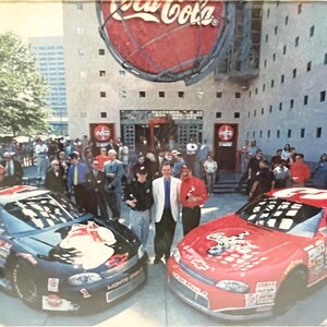 May include: Two stock cars, one black and one red, are parked in front of a Coca-Cola building. The black car has the number 1, and the red car has the number 3. A crowd of people stands in the background.