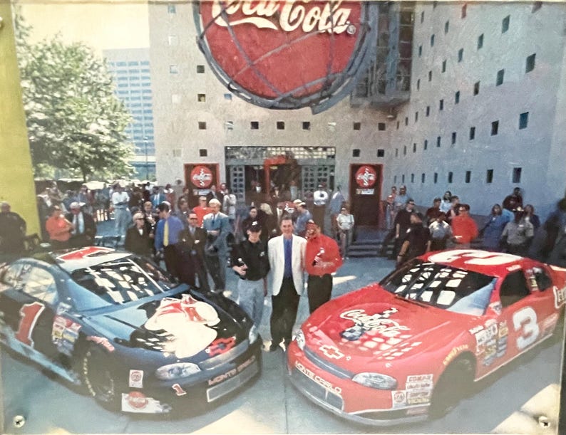 May include: Two stock cars, one black and one red, are parked in front of a Coca-Cola building. The black car has the number 1, and the red car has the number 3. A crowd of people stands in the background.
