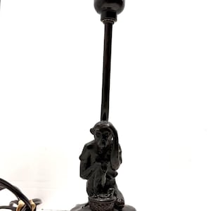 Puede incluir: Lámpara de mesa marrón oscuro con base de figura de mono. La lámpara tiene un tallo largo y delgado y un portalámparas negro. El mono está sentado, sosteniendo su cabeza, y una pequeña cesta está a sus pies. La base es redonda.