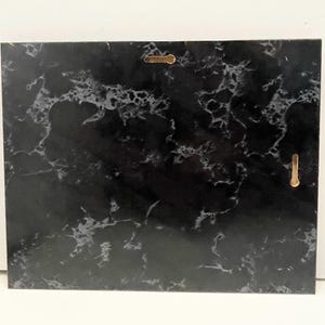 May include: A rectangular black marble plaque with white veining. Two gold-colored metal hangers are attached to the back for wall mounting. The plaque has a polished finish and a natural stone appearance.