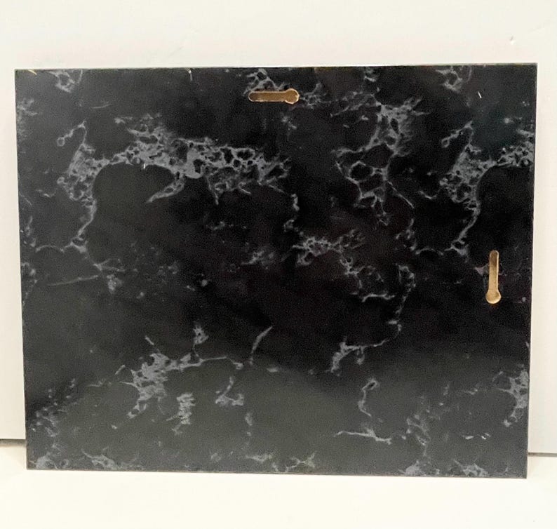 May include: A rectangular black marble plaque with white veining. Two gold-colored metal hangers are attached to the back for wall mounting. The plaque has a polished finish and a natural stone appearance.