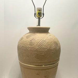 May include: Tan ceramic table lamp with a textured surface and etched swirl and line patterns. The lamp has a gold-coloured band around the middle and a black lamp shade holder.