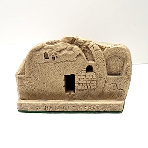 Puede incluir: Escultura de piedra beige de una tumba con una puerta abierta y las palabras "HE IS NOT HERE" en la base. La tumba tiene una superficie texturizada con detalles arquitectónicos, incluyendo una pequeña ventana y una abertura arqueada.