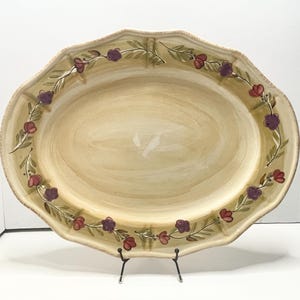 May include: Oval serving platter with a scalloped edge. The beige-colored platter features a hand-painted floral design with purple and red flowers and green leaves. The platter is supported by a black metal stand.