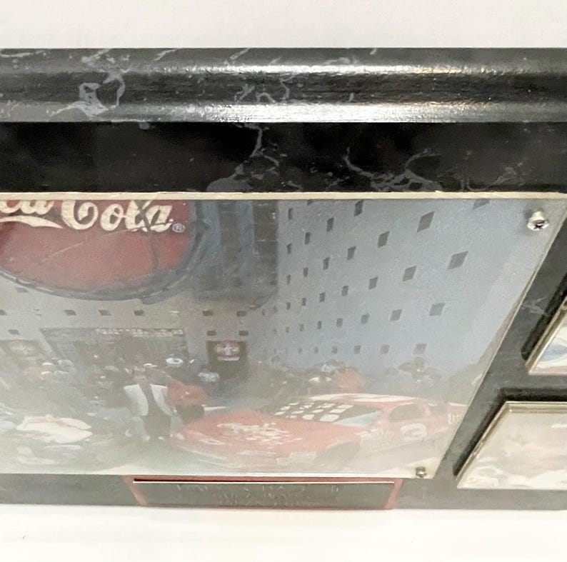 May include: A black marble-look frame with a Coca-Cola logo and a race car image. The frame has a glossy finish and a rectangular shape. The image is protected by a clear cover.
