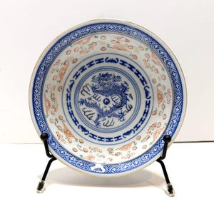 May include: A round, white porcelain plate featuring a blue dragon motif at its centre. The plate has a blue border with a repeating pattern and gold edging. It is displayed on a black metal stand.