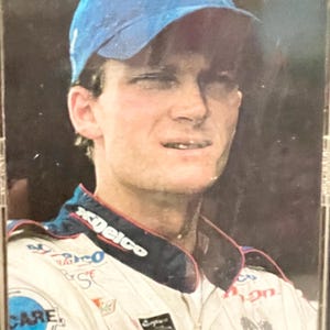 May include: A portrait of a person wearing a blue baseball cap with the number 3 on it. The person is wearing a white racing suit with sponsor logos, including "Delco" and "CARE". The image is a close-up shot.