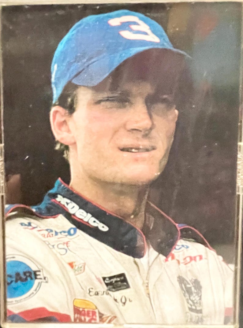 May include: A portrait of a person wearing a blue baseball cap with the number 3 on it. The person is wearing a white racing suit with sponsor logos, including "Delco" and "CARE". The image is a close-up shot.