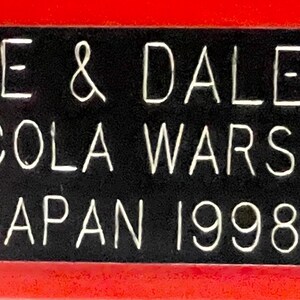 May include: A rectangular black sign with white text and a red border. The text reads "DALE & DALE JR COLA WARS JAPAN 1998". The sign is likely a commemorative item or advertisement.