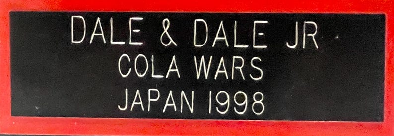 May include: A rectangular black sign with white text and a red border. The text reads "DALE & DALE JR COLA WARS JAPAN 1998". The sign is likely a commemorative item or advertisement.