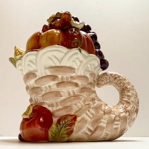 Può includere: Una cornucopia in ceramica traboccante di frutta del raccolto autunnale. Il cesto è beige e bianco, con una trama intrecciata. I frutti includono zucche, uva e una mela. Il manico è curvo e dettagliato.