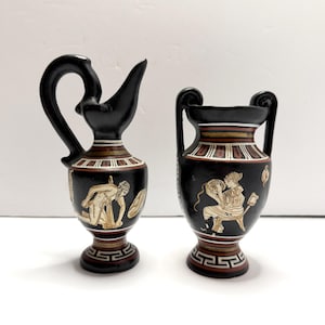 May include: Two black ceramic vessels with classical Greek-style illustrations in white and gold. The left vessel is a pitcher with a curved handle, while the right is a two-handled amphora. Both feature geometric patterns and detailed figures.