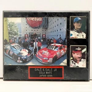 May include: A framed collage featuring two race cars and two portraits. The main image shows a Coca-Cola building with race cars in front. The text on the frame reads "DALE & DALE JR COLA WARS JAPAN 1998".