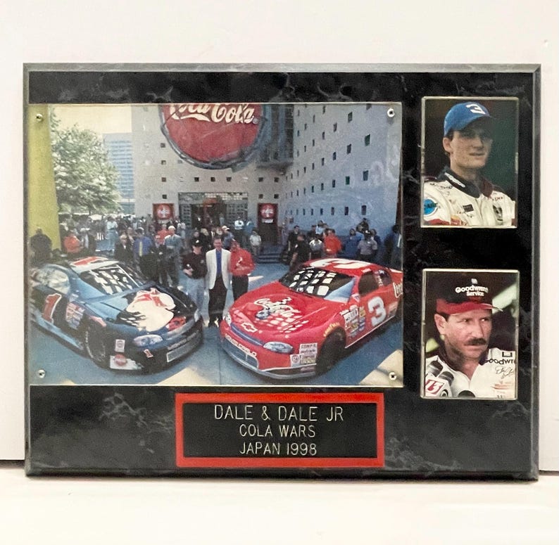 May include: A framed collage featuring two race cars and two portraits. The main image shows a Coca-Cola building with race cars in front. The text on the frame reads "DALE & DALE JR COLA WARS JAPAN 1998".