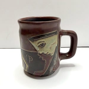 May include: A dark brown ceramic mug featuring a stylized, etched graphic of a human face in profile. The design incorporates light beige and dark charcoal tones within the etched lines, contrasting against the solid brown glaze of the mug.