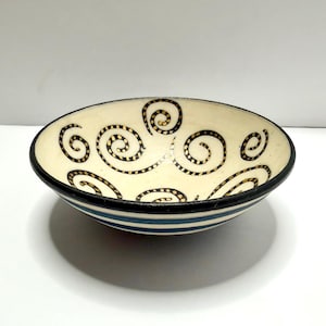May include: A shallow ceramic bowl with an off-white interior featuring black spiral patterns. The exterior of the bowl is decorated with horizontal blue stripes, and the rim is finished with a solid black band, creating a distinct contrast.