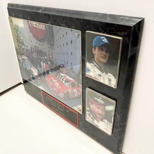 May include: A framed collage featuring racing imagery. The collage includes a large photo of a red race car with the number 3, a photo of a race car driver wearing a blue cap, and a photo of a race car driver wearing a red cap. The frame is black with a marble-like pattern.