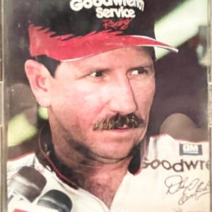 May include: Portrait of a man wearing a black and red Goodwrench Service Racing hat. He has a mustache and is wearing a white and black racing uniform. The image includes the text "Winston" and "7 Time".