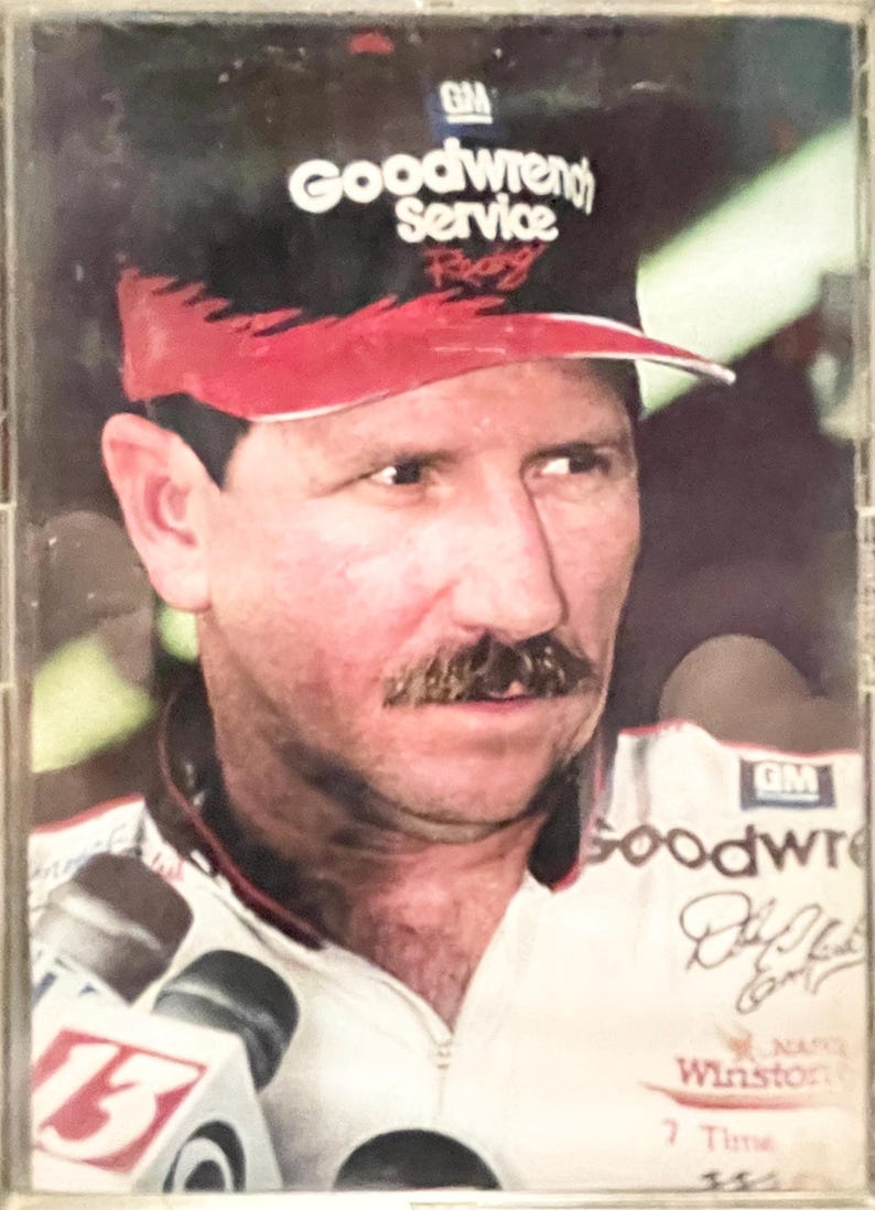 May include: Portrait of a man wearing a black and red Goodwrench Service Racing hat. He has a mustache and is wearing a white and black racing uniform. The image includes the text "Winston" and "7 Time".