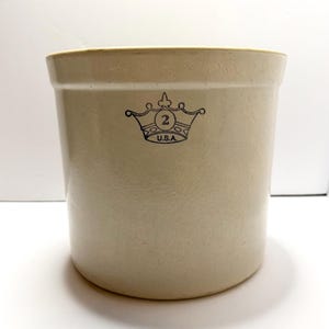 以下が含まれることがあります： 厚い縁を持つ円筒形のオフホワイトのストーンウェア製クロック。前面には、数字の2とその下にU.S.A.という文字が入った王冠の黒いスタンプグラフィックが施されています。表面は滑らかで、均一なベージュトーンの釉薬仕上げになっています。