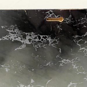 May include: A rectangular decorative panel with a black and white marble design. The panel has a keyhole-shaped hanging slot on the back for easy mounting. The marble pattern features swirling veins and a glossy finish.