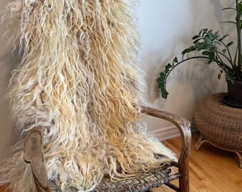 May include: A vintage wooden rocking chair with a woven seat and back. A cream-colored, shaggy sheepskin throw is draped over the back of the chair. The chair is positioned near a wall and a potted plant.